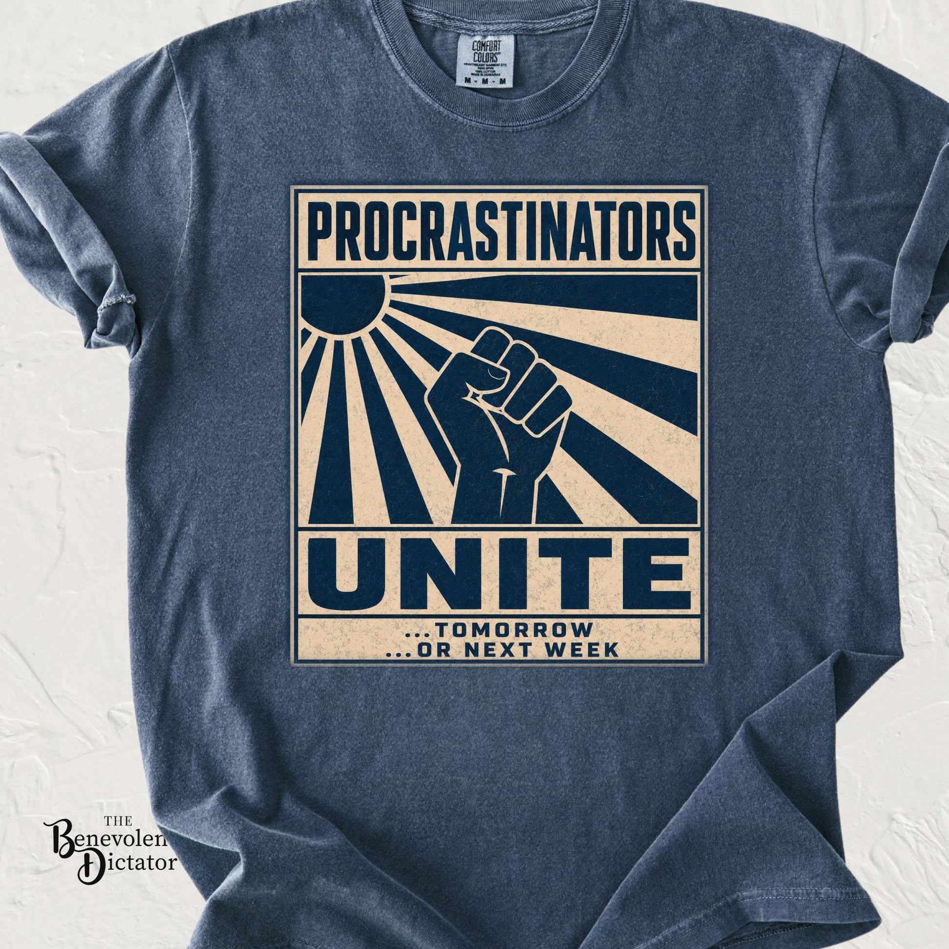 Blue Jean Comfort Colors t-shirt featuring a distressed retro-style graphic with bold cream and navy typography reading “Procrastinators Unite… Tomorrow or Next Week” with a raised fist and sunburst design.