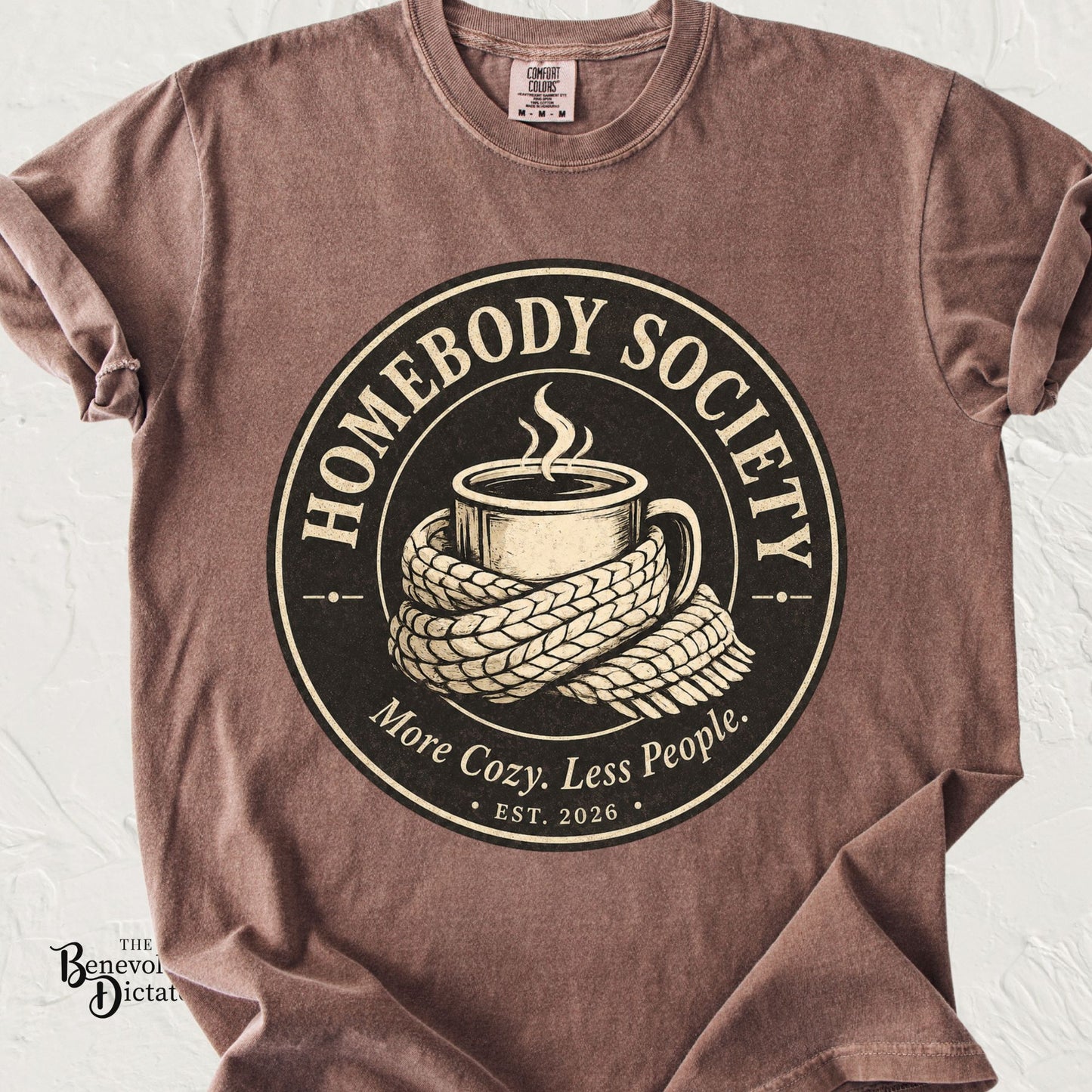 Vintage-inspired Homebody Society emblem featuring a steaming coffee mug wrapped in a cozy knit scarf with the words "More Cozy. Less People."