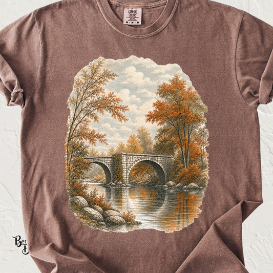 Espresso Comfort Colors t-shirt featuring a vintage autumn landscape with a stone arch bridge over calm water, surrounded by orange fall trees and foliage.
