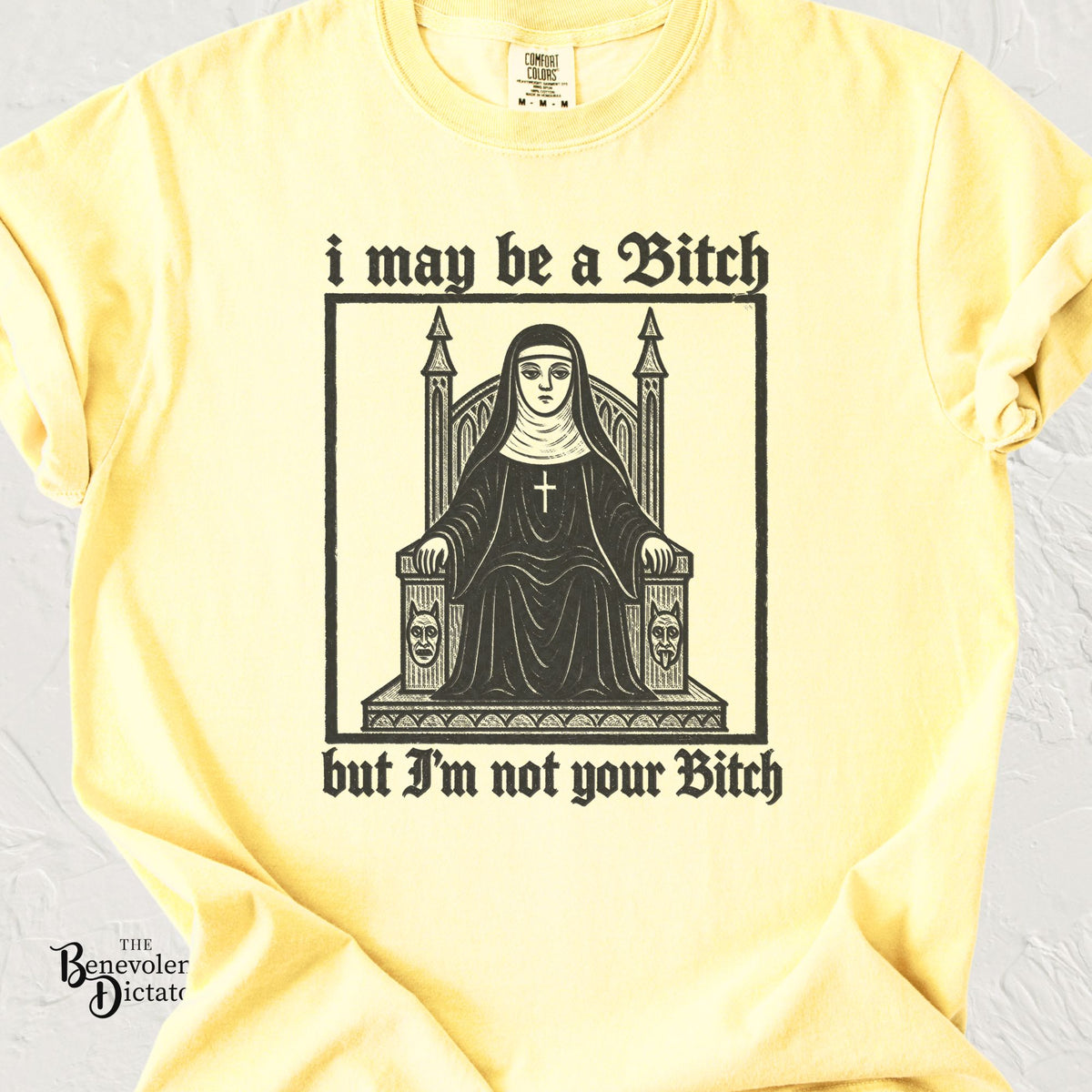 A pale yellow graphic t-shirt laid flat on a light textured background. The shirt features a black gothic-style illustration of a nun sitting on a throne with the text 'i may be a Bitch but I'm not your Bitch' printed around the graphic.