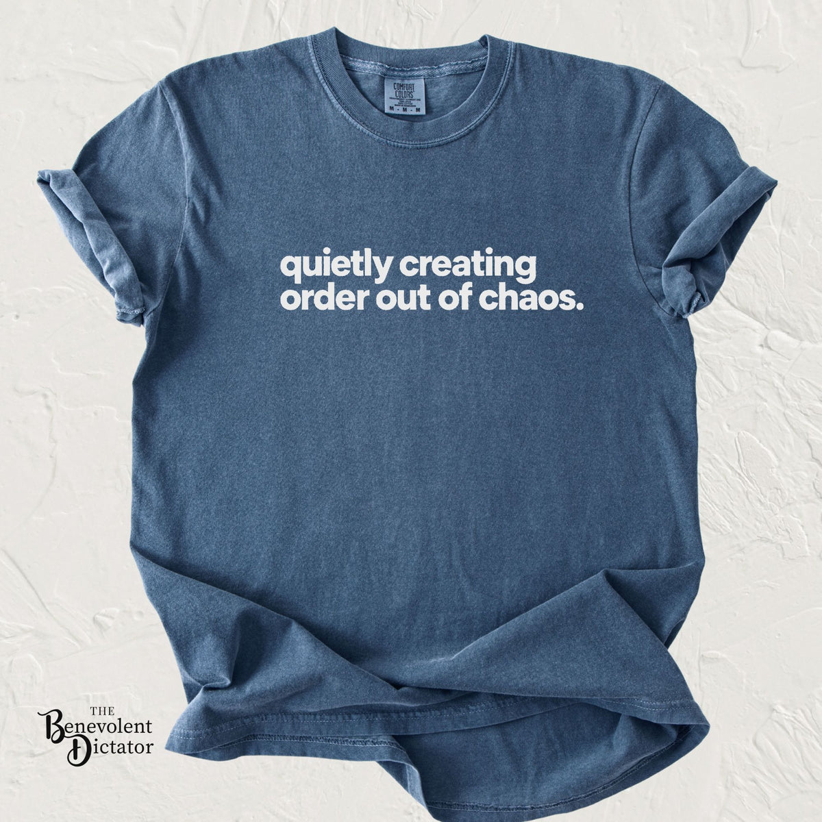 A washed slate blue t-shirt with white text reading 'quietly creating order out of chaos.' laid flat on a textured off-white background.